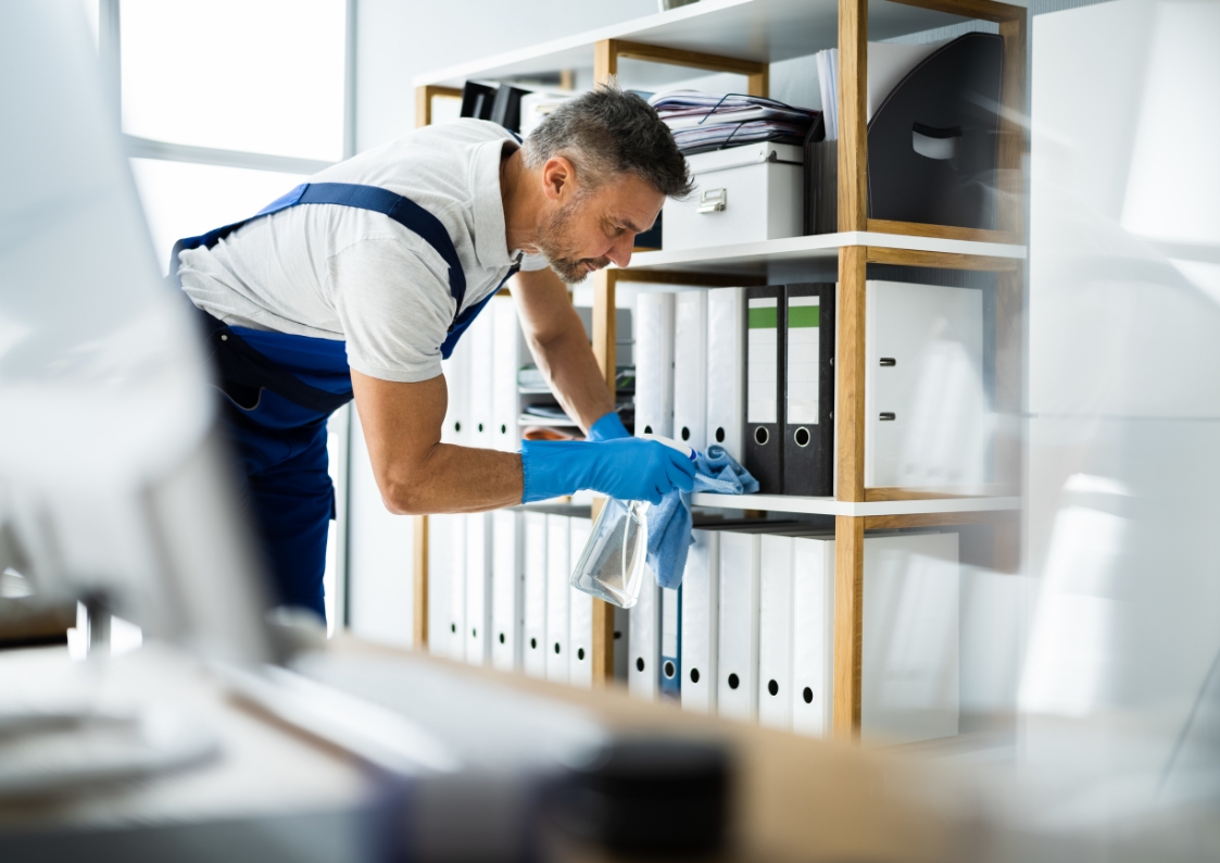 Mężczyzna myje półkę w biurze. Ma na sobie rękawiczki i trzyma w dłoniach szmatkę. Jest skupiony na pracy.