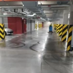 Widok garażu podziemnego podczas profesjonalnego sprzątania, z widocznymi śladami opon i wilgotną posadzką. Pracownik firmy sprzątającej obsługuje maszynę czyszczącą, usuwając zabrudzenia z betonowej podłogi. Kolumny garażowe oznaczone są czarno-żółtymi pasami ostrzegawczymi, a w tle widać zaparkowany samochód. Otoczenie jest intensywnie myte, aby zapewnić estetykę oraz bezpieczeństwo użytkownikom parkingu.