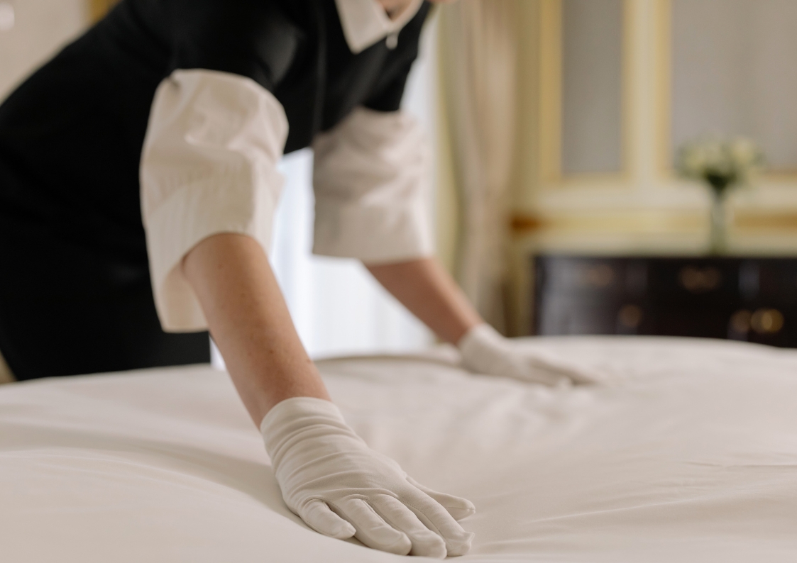 Kobieta ścieli łóżko hotelowe. Ma na sobie białe rękawiczki.